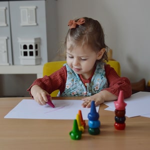 May include: A set of stackable, cone-shaped crayons in various colors including green, yellow, blue, and red, arranged on a wooden table. The crayons feature a rounded base and a pointed top, designed to be stacked vertically on top of one another.