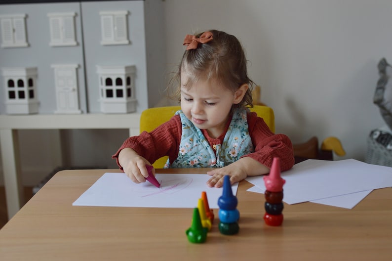 May include: A set of stackable, cone-shaped crayons in various colors including green, yellow, blue, and red, arranged on a wooden table. The crayons feature a rounded base and a pointed top, designed to be stacked vertically on top of one another.
