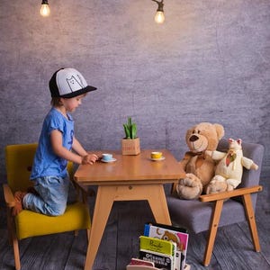 Peut inclure: Sc&egrave;ne de go&ucirc;ter pour enfants avec une petite table en bois, des chaises jaunes et grises, et des animaux en peluche. Un enfant portant une casquette avec des silhouettes de chats est agenouill&eacute; &agrave; la table. Des tasses &agrave; th&eacute; miniatures et une petite bo&icirc;te en bois avec des livres sont &eacute;galement pr&eacute;sentes.