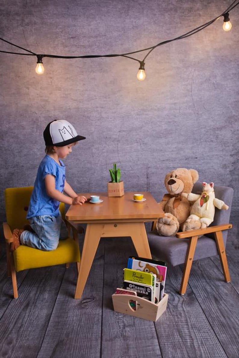 Peut inclure: Sc&egrave;ne de go&ucirc;ter pour enfants avec une petite table en bois, des chaises jaunes et grises, et des animaux en peluche. Un enfant portant une casquette avec des silhouettes de chats est agenouill&eacute; &agrave; la table. Des tasses &agrave; th&eacute; miniatures et une petite bo&icirc;te en bois avec des livres sont &eacute;galement pr&eacute;sentes.
