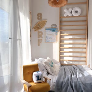 Peut inclure: Une chambre ensoleill&eacute;e sur le th&egrave;me c&ocirc;tier. Comprend un fauteuil jaune moutarde, un lit avec une couverture grise et des d&eacute;corations murales avec le mot "SURF". Un luminaire suspendu en osier est suspendu au plafond.