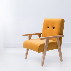 Peut inclure: Fauteuil jaune moutarde avec accoudoirs et pieds en bois. La chaise a un dossier et une assise arrondis, avec deux d&eacute;tails circulaires noirs sur le dossier. La chaise est plac&eacute;e sur un fond blanc.