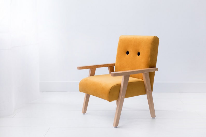 Peut inclure: Fauteuil jaune moutarde avec accoudoirs et pieds en bois. La chaise a un dossier et une assise arrondis, avec deux d&eacute;tails circulaires noirs sur le dossier. La chaise est plac&eacute;e sur un fond blanc.
