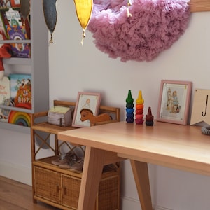 May include: A light-colored wooden desk with angled legs, featuring two stacks of colorful, tiered stacking crayons on the surface. A small rattan shelf unit sits nearby, holding a wooden horse figurine and two framed illustrations in pink frames.