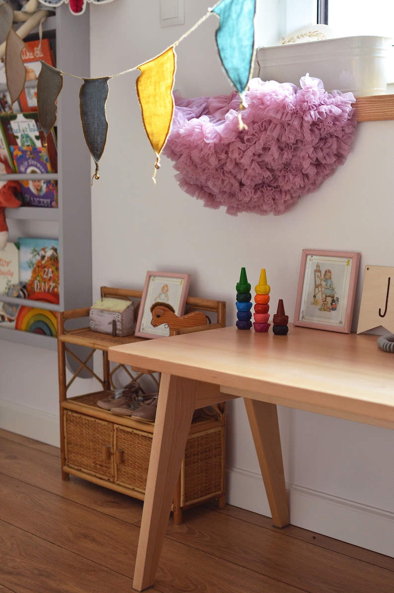 May include: A light-colored wooden desk with angled legs, featuring two stacks of colorful, tiered stacking crayons on the surface. A small rattan shelf unit sits nearby, holding a wooden horse figurine and two framed illustrations in pink frames.