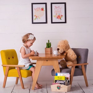 Peut inclure: Une aire de jeux pour enfants avec une petite table et des chaises en bois. Un ours en peluche est assis &agrave; la table avec une tasse et une soucoupe. Deux tableaux encadr&eacute;s sont accroch&eacute;s au mur blanc. Une petite bo&icirc;te en bois contient des livres.