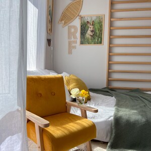 Peut inclure: Un fauteuil jaune moutarde avec des accoudoirs et des pieds en bois dans une pi&egrave;ce ensoleill&eacute;e. Une impression encadr&eacute;e de lapin et une d&eacute;coration de planche de surf en osier ornent le mur. Un lit avec une couverture verte et un oreiller d&eacute;coratif est en arri&egrave;re-plan.