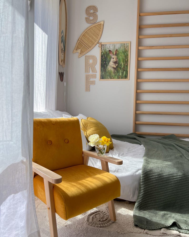 Peut inclure: Un fauteuil jaune moutarde avec des accoudoirs et des pieds en bois dans une pi&egrave;ce ensoleill&eacute;e. Une impression encadr&eacute;e de lapin et une d&eacute;coration de planche de surf en osier ornent le mur. Un lit avec une couverture verte et un oreiller d&eacute;coratif est en arri&egrave;re-plan.