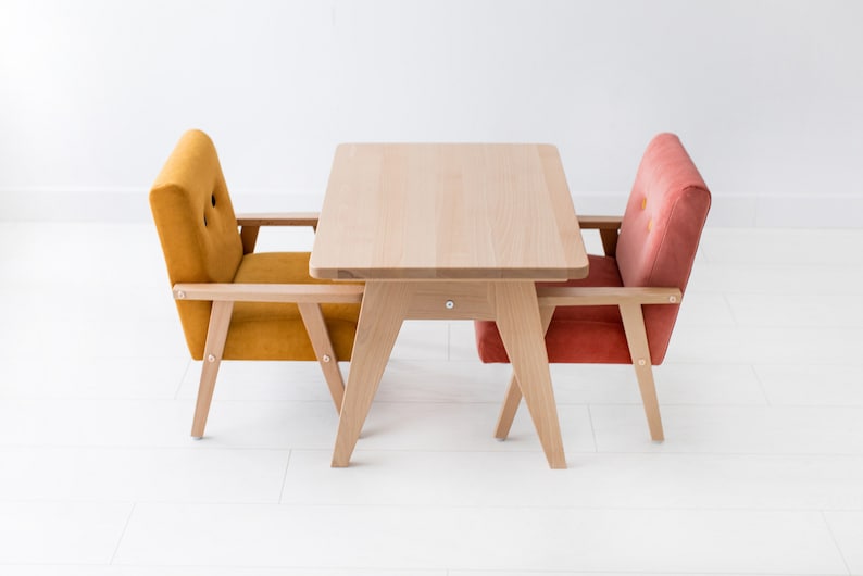 May include: A light-colored wooden table with angled legs paired with two upholstered armchairs. One chair is mustard yellow with two buttons on the backrest, and the other is a muted coral pink with two matching buttons on the backrest.