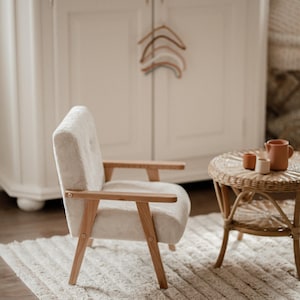 May include: A cream-colored armchair with wooden armrests and legs sits on a white rug. A small, round wicker table holds a terracotta pitcher and two small cups. A white cabinet and wooden hangers are in the background.