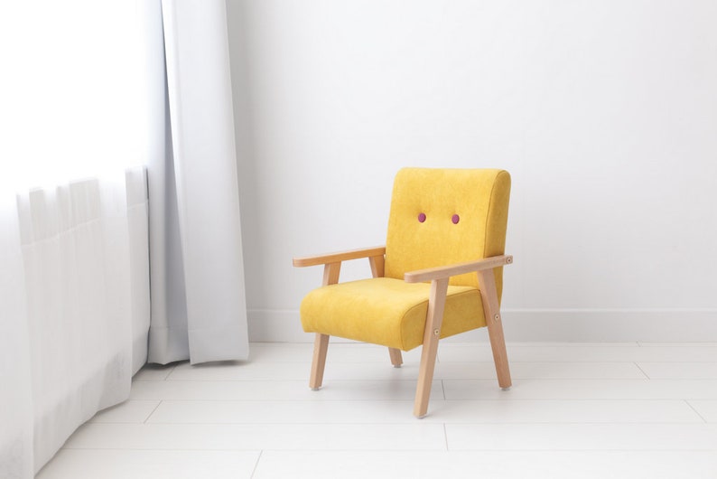 Peut inclure: Un petit fauteuil rembourr&eacute; jaune avec des accoudoirs et des pieds en bois clair. La chaise a un dossier arrondi et deux petits boutons ronds violets. La chaise est plac&eacute;e contre un mur et un sol blancs.