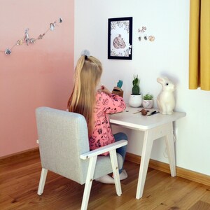 May include: A light gray upholstered armchair with white wooden legs and armrests, paired with a matching white wooden desk. The desk surface holds a white rabbit figurine, two small potted cacti, and various craft supplies on a wooden floor.