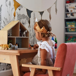 May include: A wooden dollhouse with multiple compartments containing various plastic animal figurines, placed on a light wood table. A gold sequined crown rests on the head of a child sitting in a pink velvet armchair, playing with the toys.