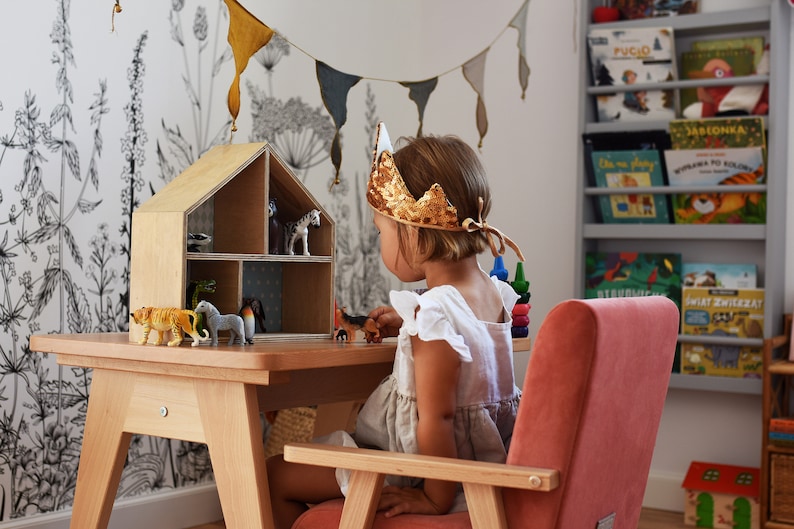 May include: A wooden dollhouse with multiple compartments containing various plastic animal figurines, placed on a light wood table. A gold sequined crown rests on the head of a child sitting in a pink velvet armchair, playing with the toys.