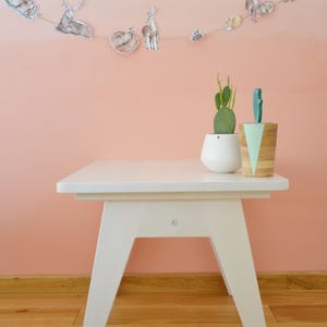 May include: A white wooden side table with angled legs, topped with a white ceramic planter containing a green cactus and a cylindrical wooden container with a mint green triangular accent, featuring a string of woodland animal illustrations.