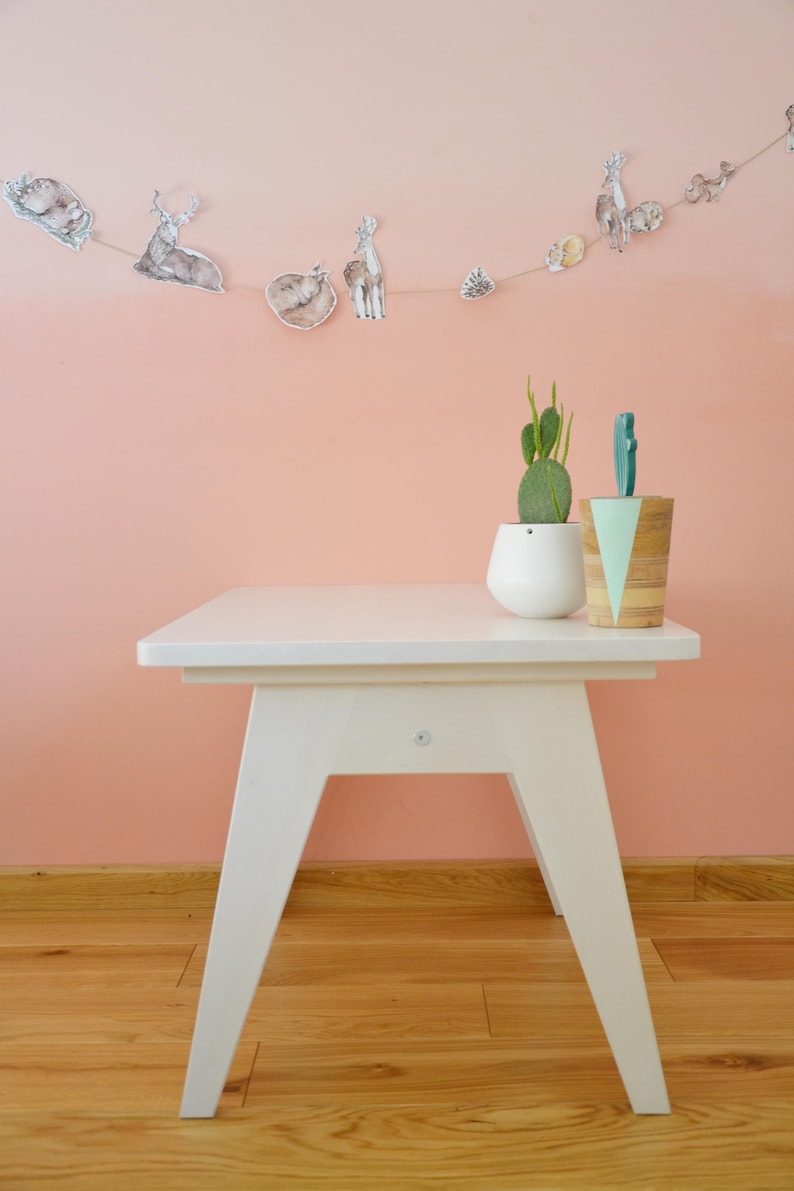 May include: A white wooden side table with angled legs, topped with a white ceramic planter containing a green cactus and a cylindrical wooden container with a mint green triangular accent, featuring a string of woodland animal illustrations.