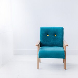 Peut inclure: Un fauteuil turquoise avec des accoudoirs et des pieds en bois. Le dossier du fauteuil est capitonn&eacute; avec deux boutons jaunes. Le fauteuil est plac&eacute; contre un mur et un sol blancs, avec un rideau blanc transparent visible &agrave; gauche.