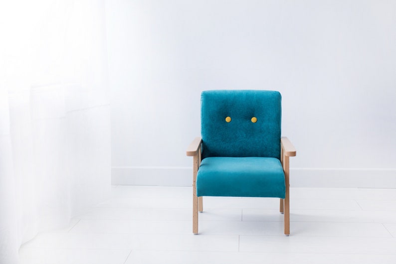 Peut inclure: Un fauteuil turquoise avec des accoudoirs et des pieds en bois. Le dossier du fauteuil est capitonn&eacute; avec deux boutons jaunes. Le fauteuil est plac&eacute; contre un mur et un sol blancs, avec un rideau blanc transparent visible &agrave; gauche.