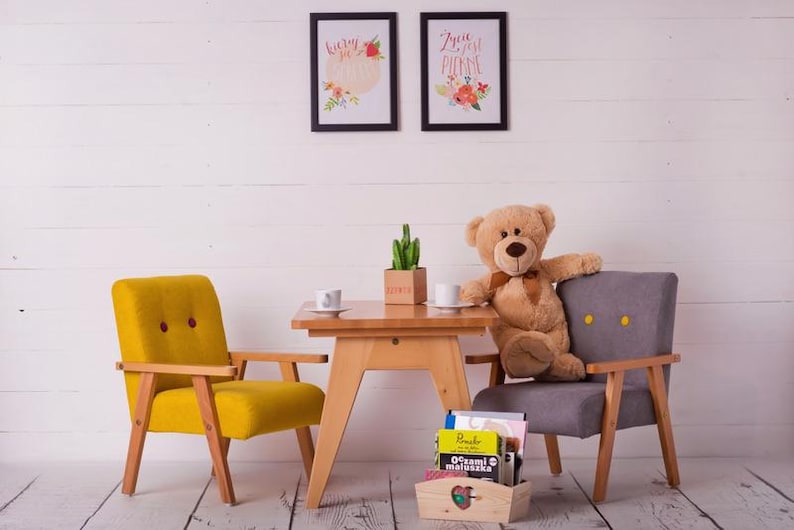 Peut inclure: Sc&egrave;ne de chambre d'enfant avec une petite table en bois, deux fauteuils et un ours en peluche. Le fauteuil jaune a deux boutons rouges. Le fauteuil gris a deux boutons jaunes. Deux tableaux encadr&eacute;s sont accroch&eacute;s au mur blanc.