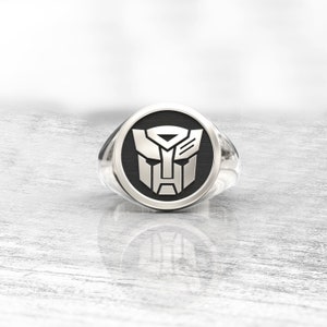 Könnte beinhalten: Ein silberner Ring mit schwarzem Gesicht, der das Autobot-Symbol zeigt. Der Ring hat eine polierte Oberfläche und ein abgerundetes Band. Das Autobot-Symbol ist ein stilisierter Roboterkopf mit geometrischen Formen.