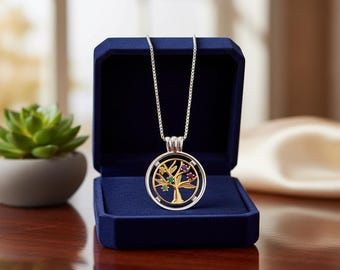 May include: A silver necklace with a gold tree pendant, set with colorful gemstones, rests in a dark blue velvet jewelry box. The tree pendant has green, purple, and yellow accents. The necklace is displayed on a dark wooden surface.