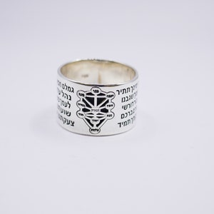 Puede incluir: Un anillo de plata con un diseño negro del Árbol de la Vida y texto hebreo alrededor de la banda.