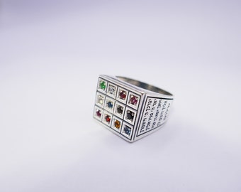 Op de afbeelding: Een zilveren ring met een vierkant oppervlak, met een raster van kleurrijke edelstenen. De ring heeft Hebreeuwse tekst gegraveerd op de zijkant. De edelstenen zijn in verschillende kleuren, waaronder groen, wit, rood, blauw en oranje.