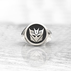 Könnte beinhalten: Ein silberner Ring mit schwarzem Gesicht, der ein silbernes Emblem zeigt. Das Emblem ist ein stilisiertes, geometrisches Design mit scharfen Winkeln und einer zentralen V-Form. Der Ring hat eine polierte, reflektierende Oberfläche.
