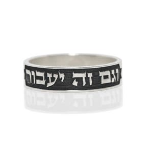 May include: A silver ring with a dark band featuring Hebrew text. The ring is displayed on a white background, with a reflection visible below. The text is in a contrasting color, making it stand out.