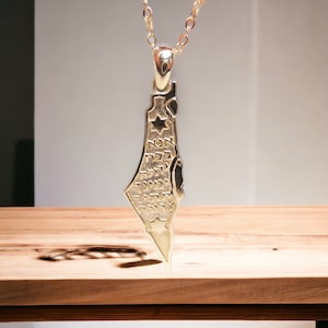 Puede incluir: Un collar con colgante dorado con un contorno de mapa detallado. El colgante tiene un diseño de estrella y texto hebreo. El collar cuelga de una delicada cadena dorada, sobre un fondo neutro.