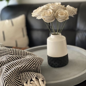 May include: A bouquet of paper roses made from book pages in a white and black vase on a gray concrete tray. The roses are arranged in a cluster and have a vintage look. The vase has a textured black bottom and a white top.