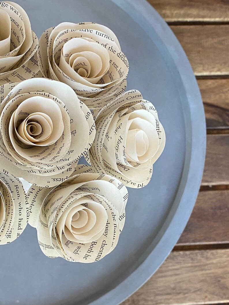May include: A bouquet of four paper roses made from the pages of a book. The roses are arranged in a circle on a gray plate. The roses are a light cream color and have a delicate, vintage look.