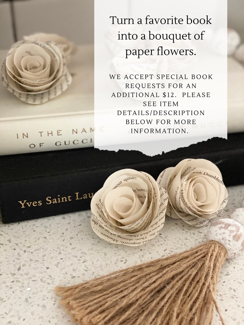 May include: Two paper flowers made from book pages, one white and one cream, are arranged on a black book with the text "Yves Saint Laurent" on the cover. The flowers are on a white surface with a brown tassel.