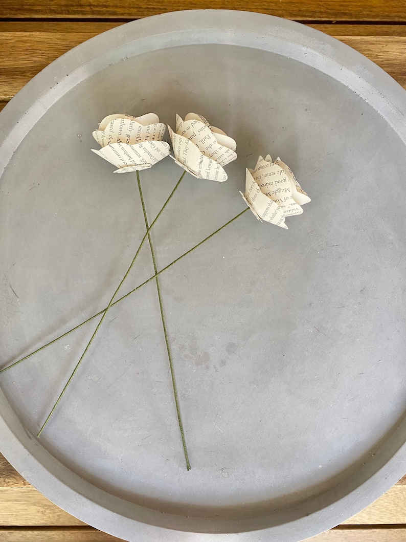 May include: Three paper roses made from book pages, with stems, on a gray concrete tray.