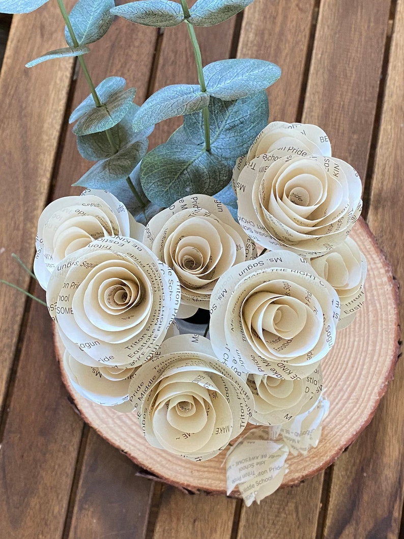 May include: A bouquet of paper roses made from book pages, arranged in a wooden planter. The roses are cream colored and have a vintage aesthetic. The planter is a natural wood slice with the bark still attached.