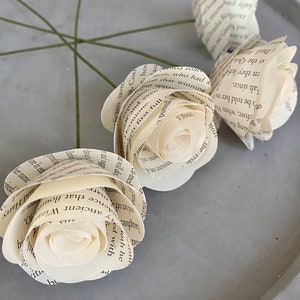 May include: Three white paper roses made from book pages with green stems. The roses are arranged on a gray surface.
