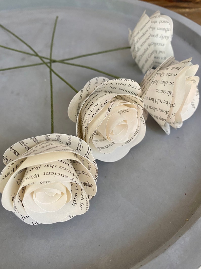 May include: Three white paper roses made from book pages with green stems. The roses are arranged on a gray surface.