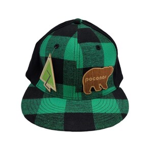May include: A black and green plaid baseball cap with a flat brim. The cap features a wooden bear patch with the word "pocono" and a tag with a tree logo. The back of the cap is black.