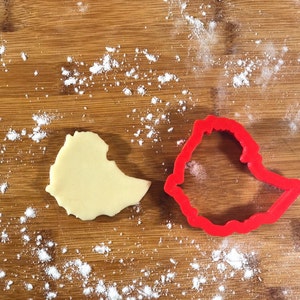 May include: A red plastic cookie cutter shaped like the outline of Ethiopia, next to a piece of cookie dough cut into the same shape on a wooden surface covered in white flour.