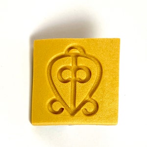 May include: A square, golden-yellow object with a textured surface. The object features a heart-shaped design with a cross and decorative swirls. The design is embossed into the surface, creating a subtle relief.
