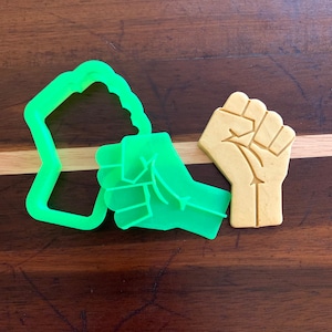 Può includere: Due stampini per biscotti in plastica verde a forma di pugno alzato. Uno stampino è pieno e l'altro è delineato.