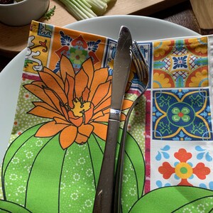 Puede incluir: Una servilleta verde y estampada doblada con un dise&ntilde;o de cactus y flores, colocada sobre un plato blanco. Un cuchillo y un tenedor de acero inoxidable est&aacute;n sobre la servilleta. Aceitunas, apio y un vaso de zumo de naranja est&aacute;n en el fondo.