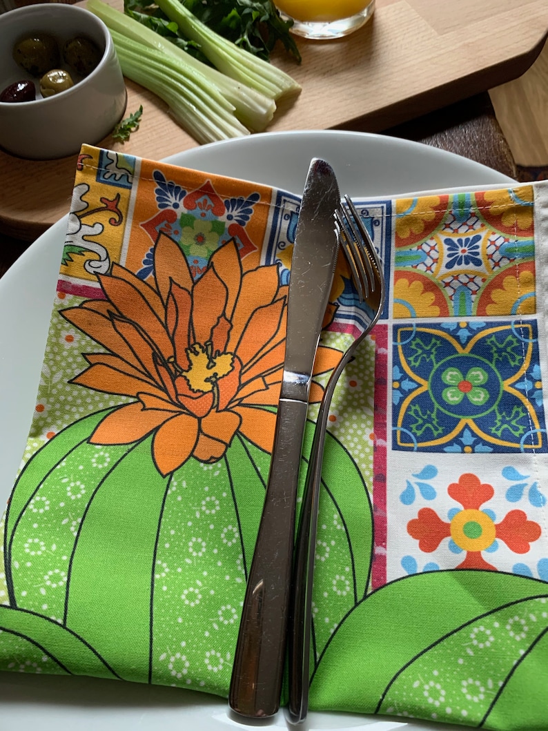 Puede incluir: Una servilleta verde y estampada doblada con un dise&ntilde;o de cactus y flores, colocada sobre un plato blanco. Un cuchillo y un tenedor de acero inoxidable est&aacute;n sobre la servilleta. Aceitunas, apio y un vaso de zumo de naranja est&aacute;n en el fondo.