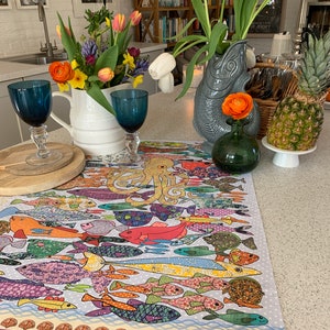 May include: A colorful table runner with a pattern of fish and an octopus. The runner is on a white countertop with a white pitcher, blue glasses, and a green vase with a yellow flower.
