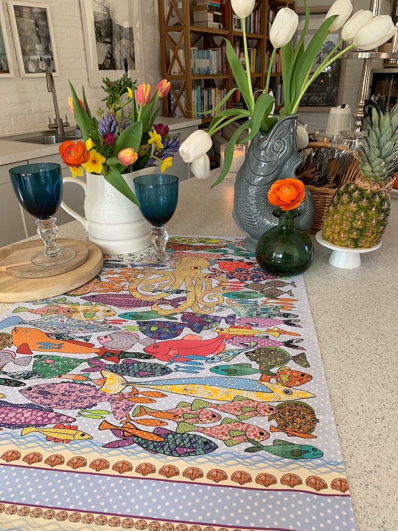 May include: A colorful table runner with a pattern of fish and an octopus. The runner is on a white countertop with a white pitcher, blue glasses, and a green vase with a yellow flower.