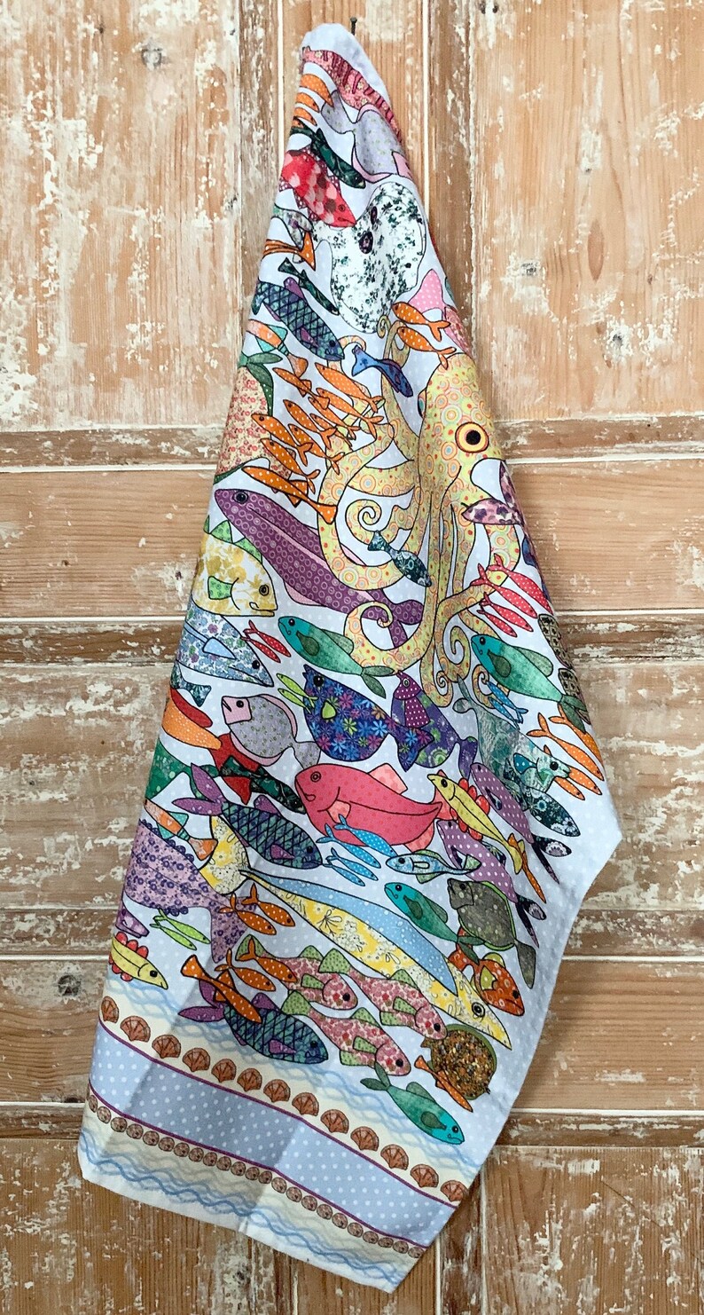 May include: A white tea towel with a colorful design of an octopus and various fish. The design is surrounded by a blue and white border with a wave pattern.