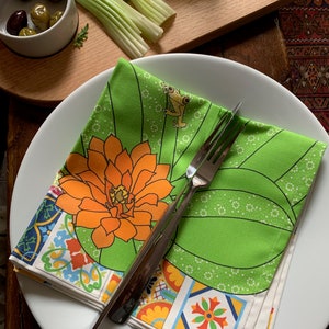 Puede incluir: Una servilleta floral verde y blanca doblada con una flor naranja y un dise&ntilde;o de rana, colocada en un plato blanco con un tenedor plateado. La servilleta tiene un borde de azulejos coloridos. Aceitunas, apio y un vaso peque&ntilde;o de l&iacute;quido en el fondo.