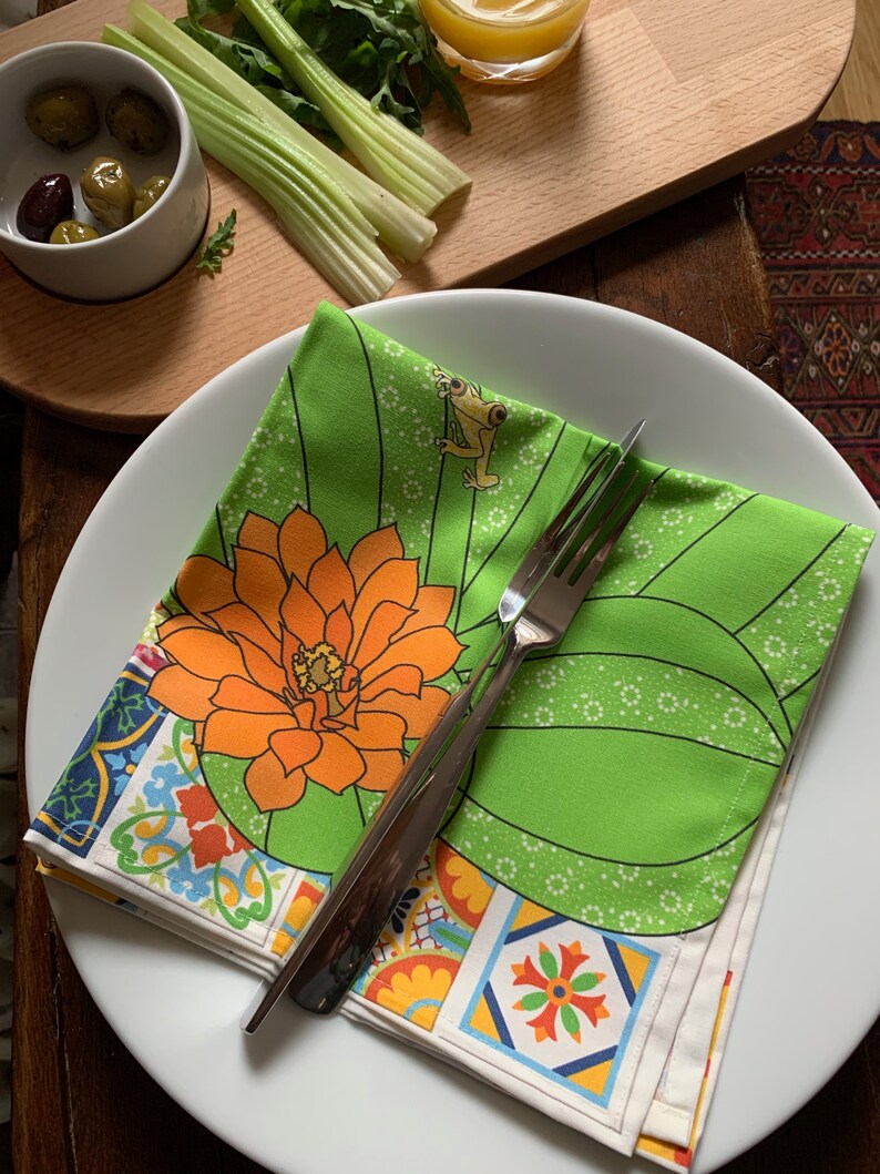 Puede incluir: Una servilleta floral verde y blanca doblada con una flor naranja y un dise&ntilde;o de rana, colocada en un plato blanco con un tenedor plateado. La servilleta tiene un borde de azulejos coloridos. Aceitunas, apio y un vaso peque&ntilde;o de l&iacute;quido en el fondo.
