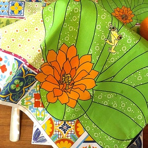 Puede incluir: Un vibrante camino de mesa y servilletas con tem&aacute;tica de cactus verde con una flor naranja y un dise&ntilde;o de rana amarilla. El camino de mesa presenta un colorido estampado de azulejos y un estampado floral. Una naranja fresca est&aacute; sobre la mesa.