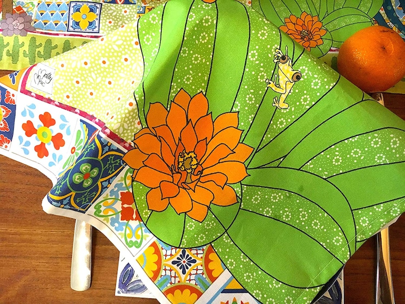 Puede incluir: Un vibrante camino de mesa y servilletas con tem&aacute;tica de cactus verde con una flor naranja y un dise&ntilde;o de rana amarilla. El camino de mesa presenta un colorido estampado de azulejos y un estampado floral. Una naranja fresca est&aacute; sobre la mesa.